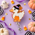 White duck badge reel wearing a black witch hat with a purple band, holding an orange pumpkin bucket, with orange glitter and star accents.