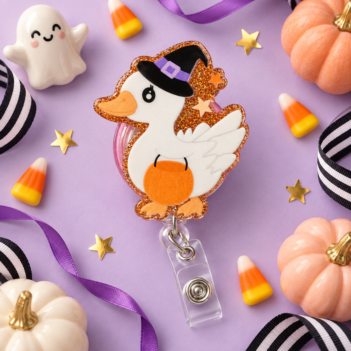 White duck badge reel wearing a black witch hat with a purple band, holding an orange pumpkin bucket, with orange glitter and star accents.