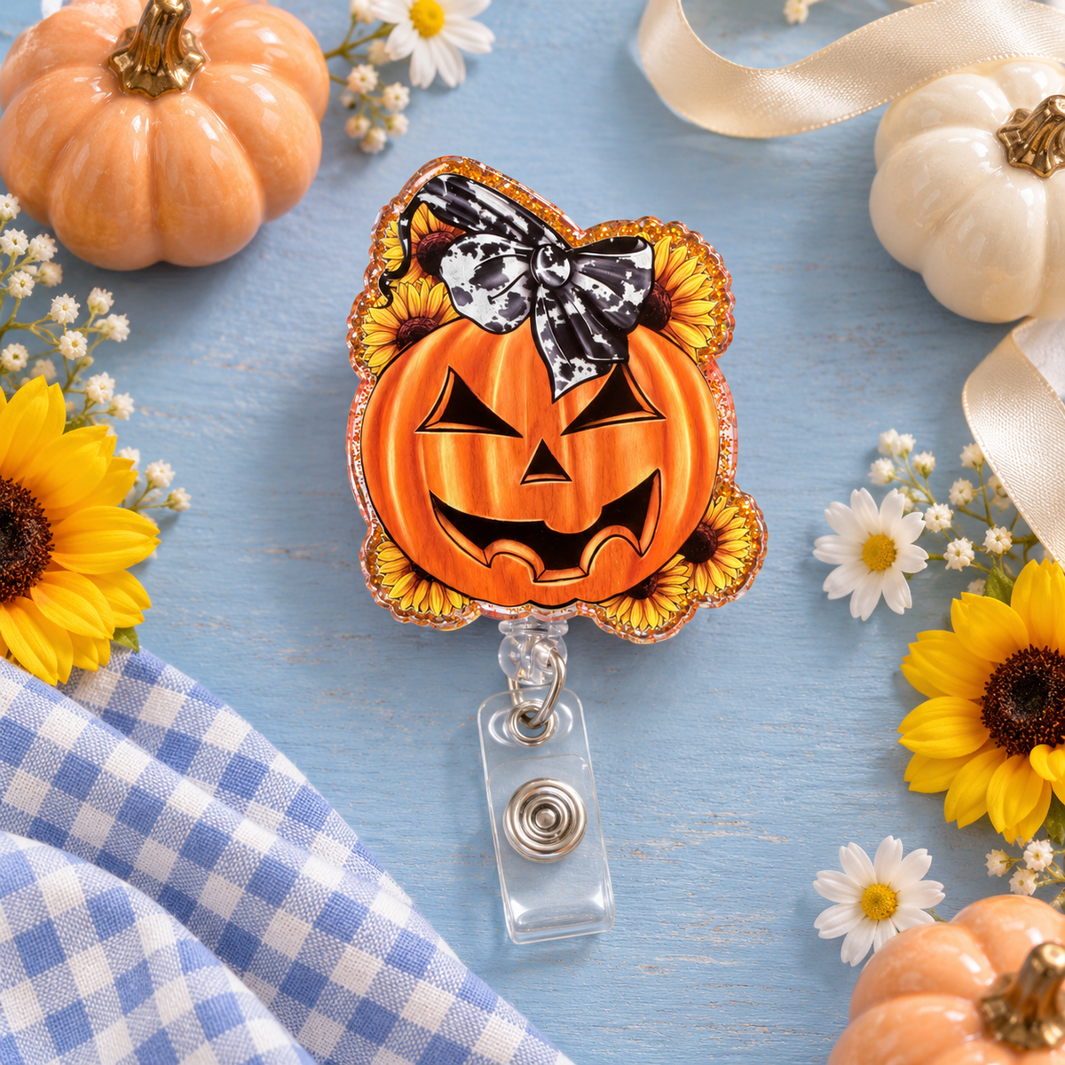 Orange jack-o’-lantern badge reel with yellow sunflowers, a black-and-white cow-print bow design, glittery orange border, and clear snap strap